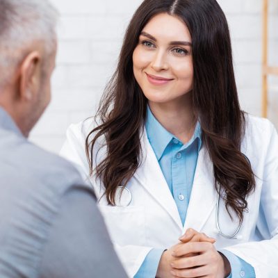 Health care, modern family doctor and medical examination. Smiling friendly young woman in white coat listens to adult patient man, sits at table in office interior, vertical, empty space, cropped